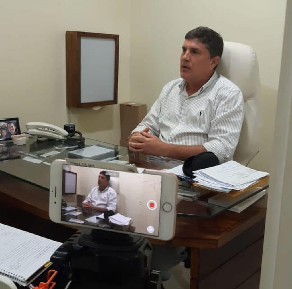 Imagem enquadra duplamente a cena (a real e a captada por uma câmera de celular deitada, que está gravando e aparece posicionada do centro para a esquerda na parte debaixo da foto). José Muniz está com camisa social branca sentado numa cadeira de escritório branca por trás de uma mesa de trabalho (com papéis, telefone, porta-retrato etc). Ele olha para a frente, como se estivesse falando com alguém. Pendurada na parede lateral, um quadro de luz para raio X com moldura de madeira, sugere que ele esteja em uma clínica odontológica.