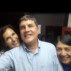 Close em trio sorridente, com enquadramento basicamente dos rostos e de parte dos ombros e da gola da camisa social azul claro do José Muniz. Da esquerda para a direita: mulher com queixo encostado sobre o ombro do José Muniz, que está ao centro, dando a impressão de que tem um braço para a frente, como se estivesse segurando o celular para uma selfie. Em seguida, com o rosto encostado na parte de cima do outro braço do dentista, rosto de outra mulher.