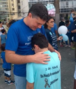 Com várias pessoas aglomeradas ao fundo, close em José Muniz (de cabeça abaixada) num abraço com menino autista (de costas), que também tem a cabeça levemente abaixada encostando no peito do dentista. O momento é de concentração para a caminhada do autismo e todos vestem camisetas azuis.