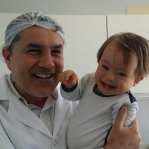 José Muniz com jaleco e touca branca sorri para foto segurando bebê especial, também sorridente, na clínica odontológica do Instituto José Muniz.