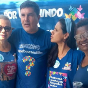 Ao ar livre, José Muniz entre três militantes femininas da causa azul posam para foto durante concentração para a caminhada de conscientização do autismo. Todos vestem camisetas azuis com programação visual da ação. Ao fundo, aparece parte de uma faixa que também faz parte do evento.