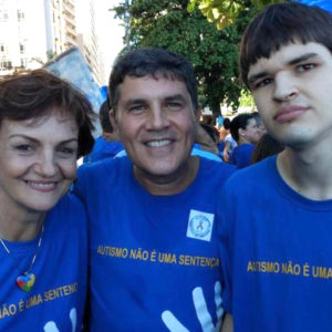 Ao ar livre, José Muniz (ao centro) abraçado com jovem especial e com Berenice Piana (a mãe do rapaz. Todos olham para a frente e usam camiseta azul com tema da caminhada de conscientização do autismo.