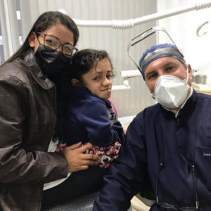 Foto no consultório da clínica odontológica do Instituto José Muniz. Do lado esquerdo, mãe de paciente especial olha para a câmera sorridente. Ela usa óculos, máscara contra covid e jaqueta de couro. Está com uma das mãos sobre sua filha especial, meio de perfil, que também olha para a câmera e veste casaco azul marinho. Do lado direito, José Muniz está de touca e jaleco azul marinho, óculos especial para o atendimento odontológico sobre a cabeça e máscara branca contra covid.