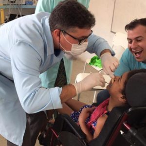 José Muniz com jaleco azul, máscara, luvas e óculos de perfil, em atendimento odontológico a menina com paralisia cerebral, que também aparece de perfil. No canto direito da foto, rosto sorridente do pai da criança.