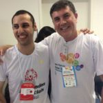 Ao fundo, paredes brancas. Em close, enquadrados da cabeça à região peitoral, jovem especial e José Muniz abraçados e sorrindo para a foto. Ambos usam camiseta branca da Special Olympics.