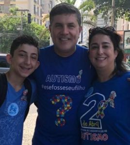 Ao ar livre, José Muniz (ao centro) abraçado com menino especial e mãe da criança. Todos olham para a frente, sorriem e usam camiseta azul com tema da caminhada de conscientização do autismo.