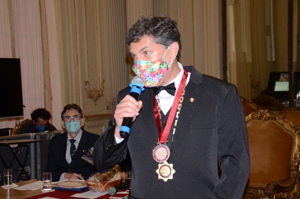 Ao fundo, Câmara Municipal do Rio de Janeiro com dois membros da Academia Brasileira de Odontologia (ABO). Em close, José Muniz em traje de passeio completo tem o microfone às mãos e usa máscara contra Covid com tema de quebra-cabeça colorido (em referência ao autismo). Enquadramento mostra rosto e tronco do dentista e, sobre a roupa, duas medalhas.